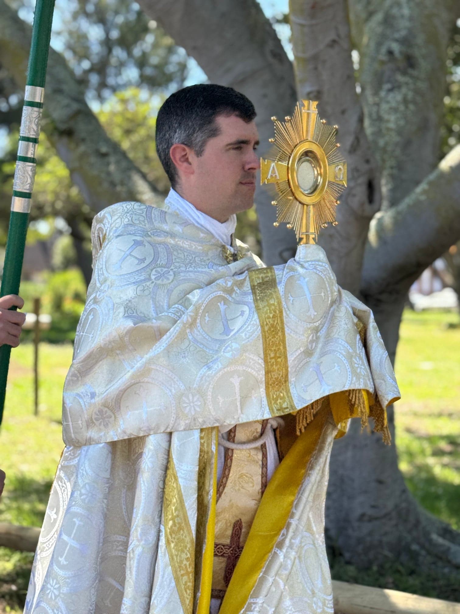 Father Keiser in white and gold vestments holding the monstrance with the Blessed Sacrament under the trees of the park, during the Christ the King procession